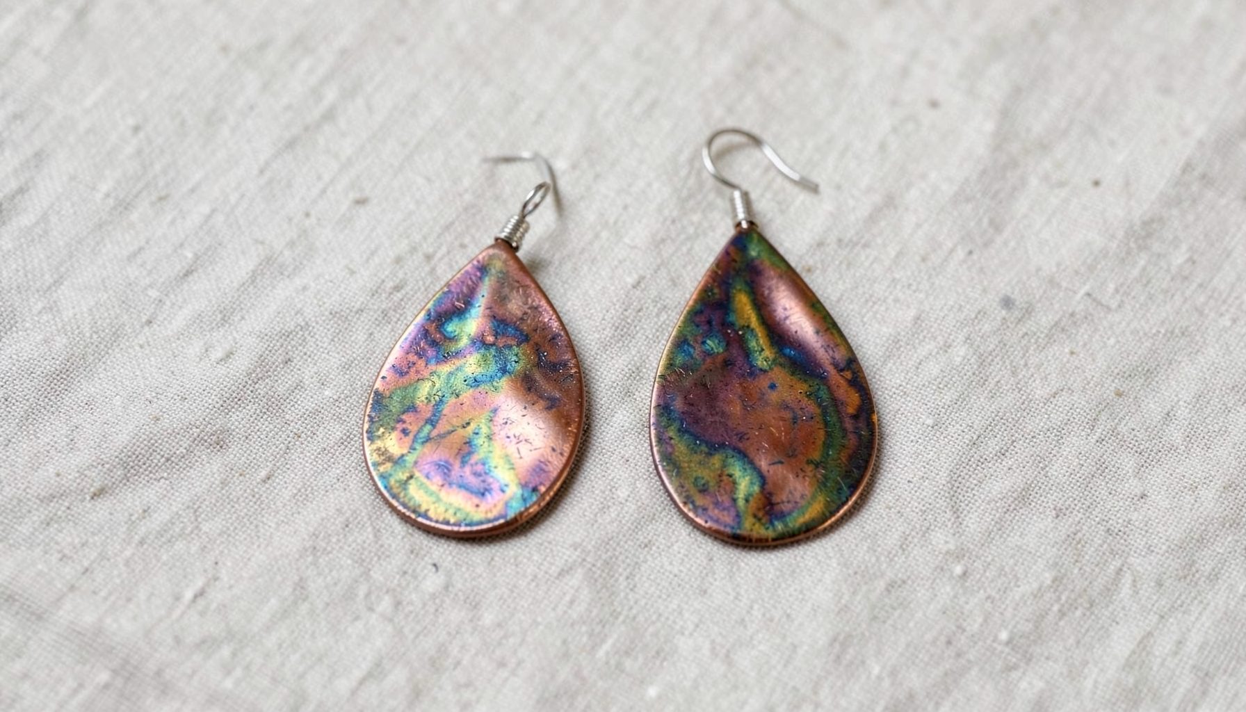 A pair of hand-formed copper paddle earrings with rich metallic glaze, resting on a neutral linen background