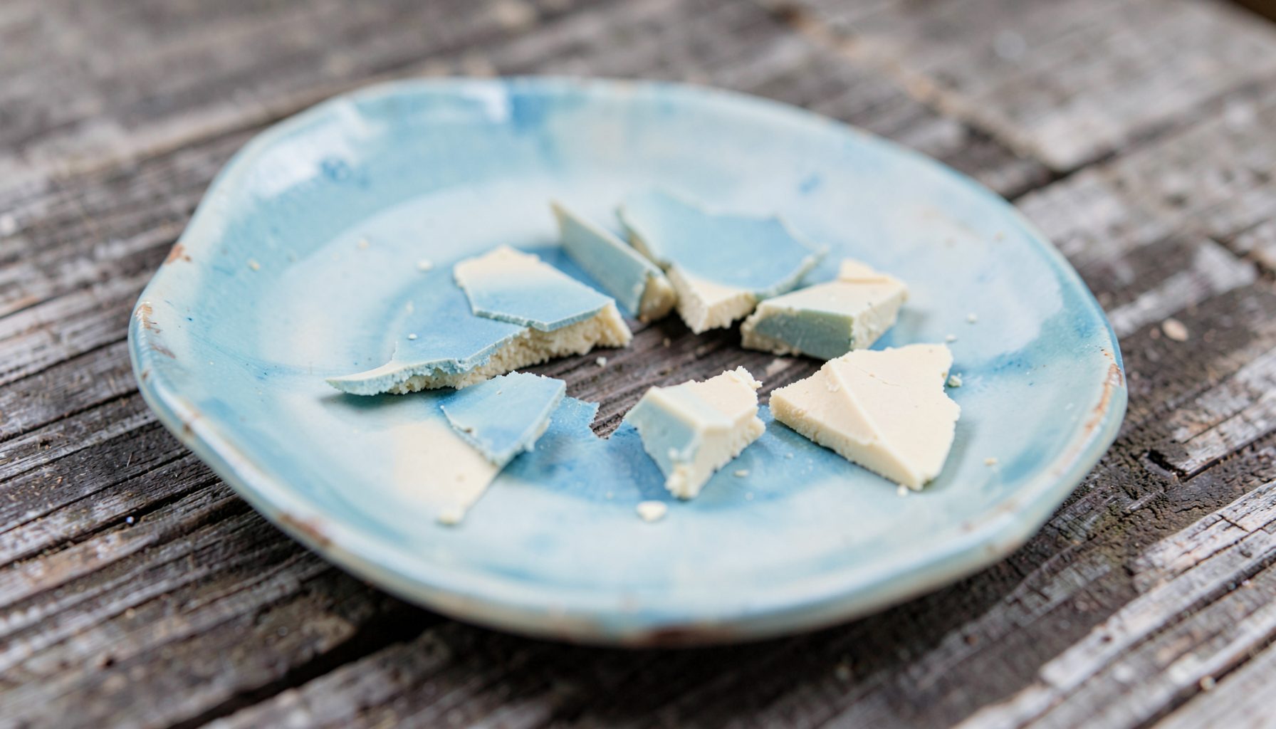 A close view of broken vintage ceramic plate shards in soft blue and cream, resting on a weathered wooden surface