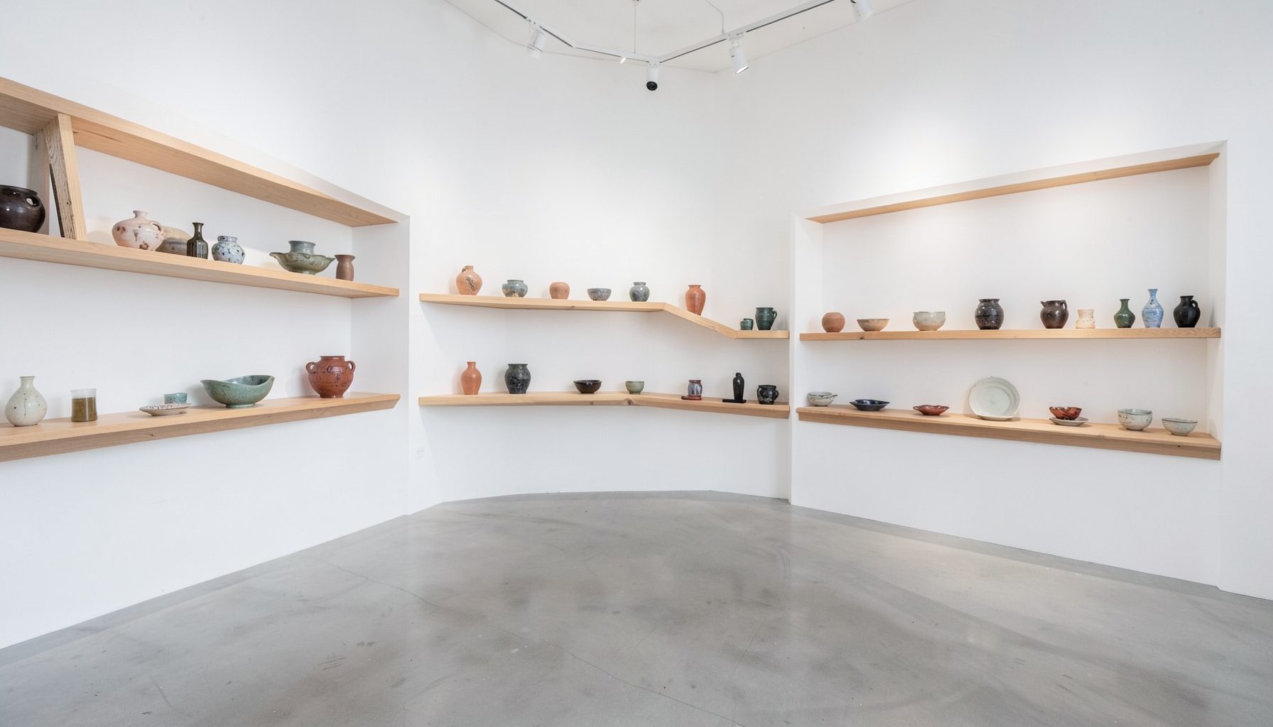 A bright gallery interior with white walls and wooden display shelves holding ceramic pieces