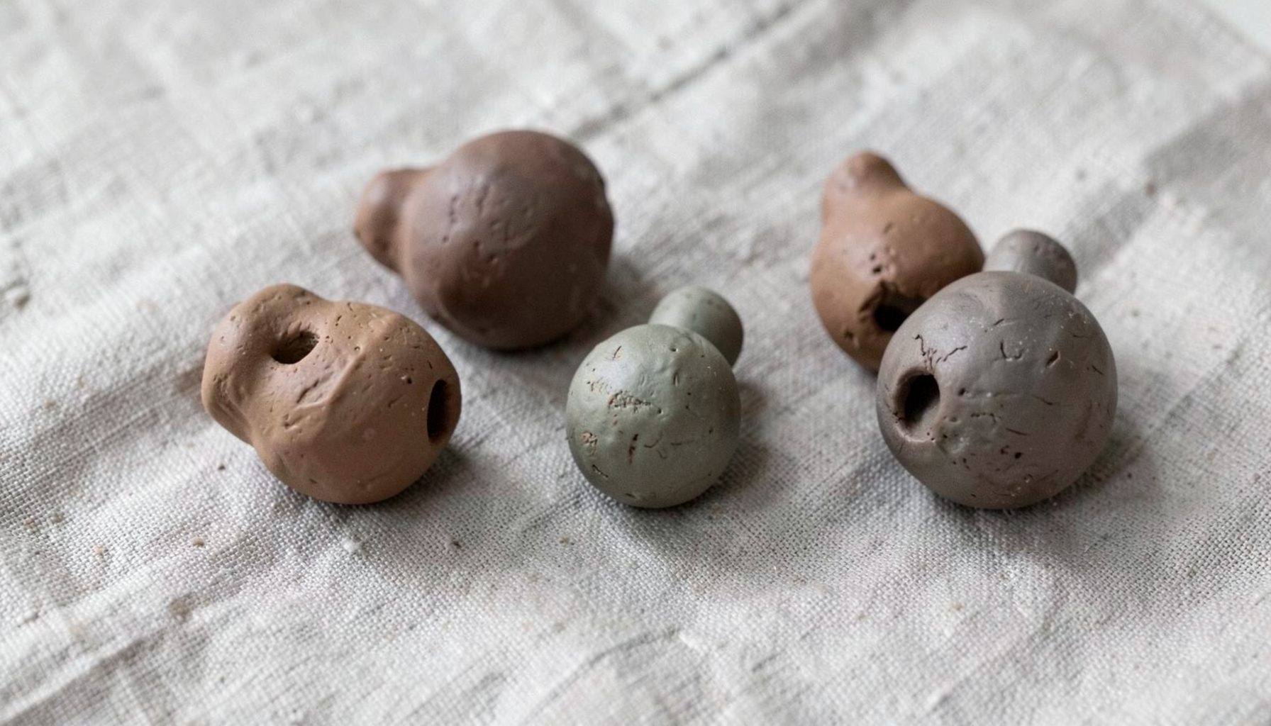 Several hand-formed clay rattles in muted earth tones arranged on a textured linen cloth