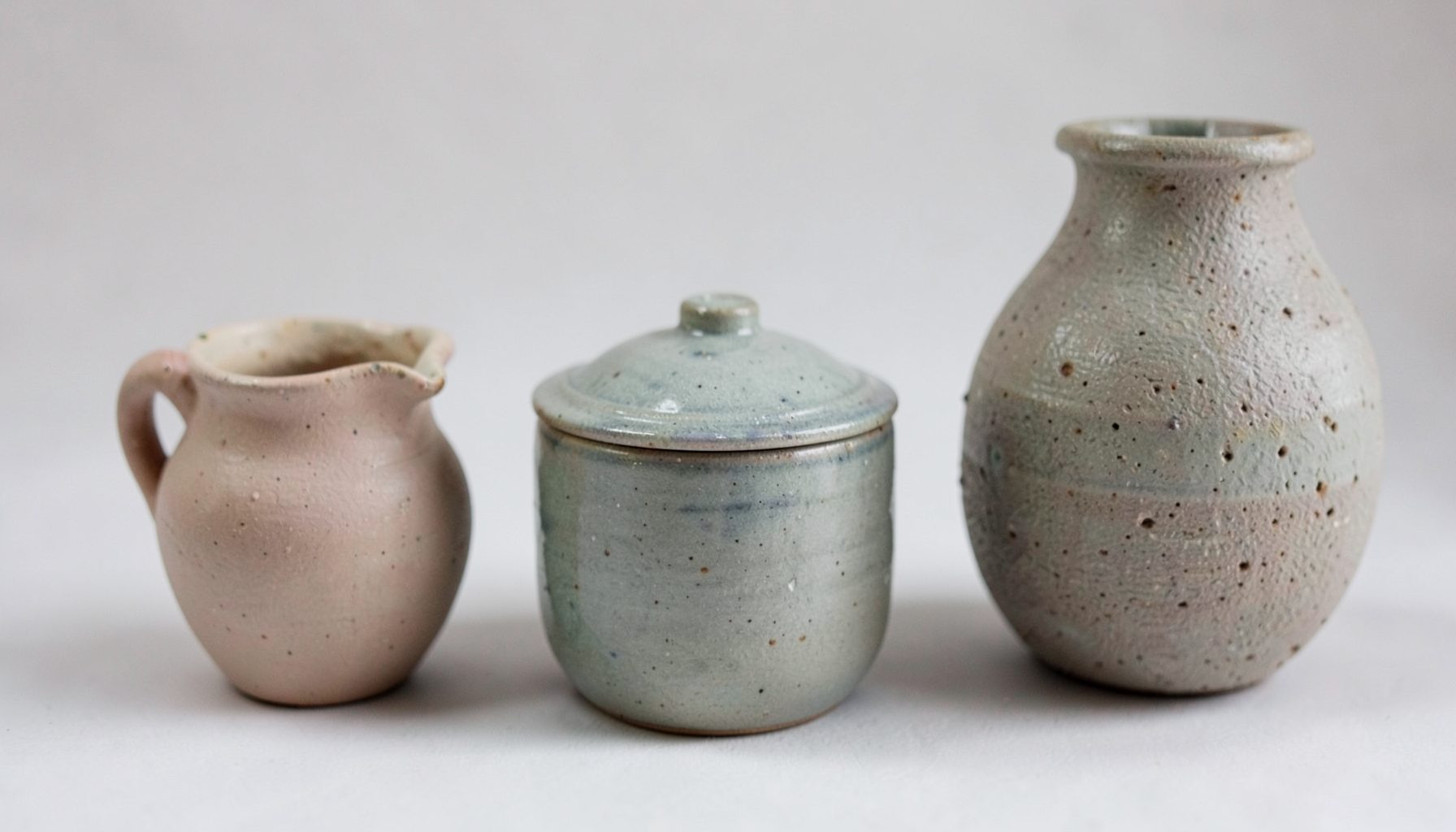Close-up of hand-built ceramic forms — a small pitcher, a lidded jar, and a textured vase in muted glazes